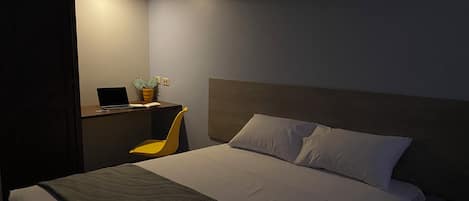 Una cama bien hecha con una manta gris, un cabecero de madera y un pequeño escritorio con una computadora portátil y una planta en maceta.