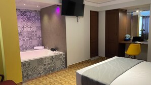 Ein Hotelzimmer mit Bett, Schreibtisch mit Laptop, gelber Stuhl, Bad mit Badewanne und an der Wand montiertem Fernseher.