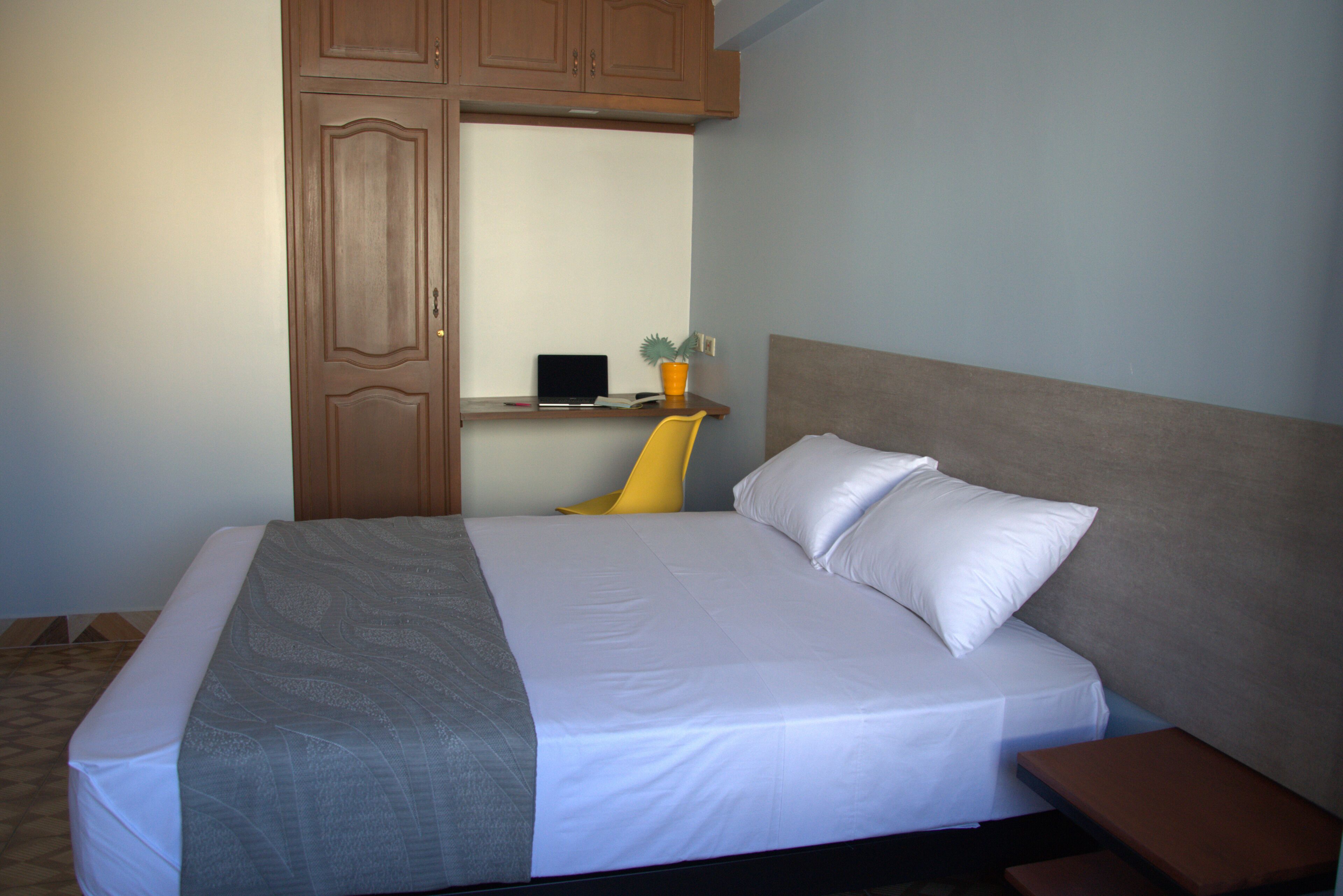 Una cama bien hecha con cabecera, una mesita de noche, una silla y un escritorio con una planta.