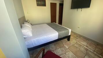 A bedroom with a bed, a TV mounted on the wall, a small window, and a stone floor.