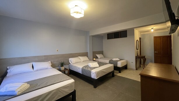 Ein Hotelzimmer mit drei nebeneinander stehenden Betten, einem an der Wand montierten Fernseher und einem kleinen Tisch mit einem Stuhl.