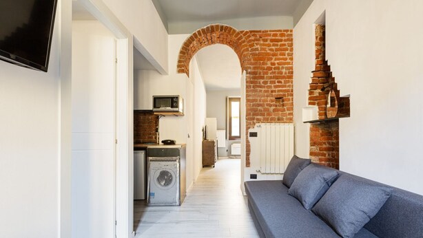 A modern living room with a grey sofa, a TV mounted on the wall, a brick fireplace, and a kitchen area in the background.