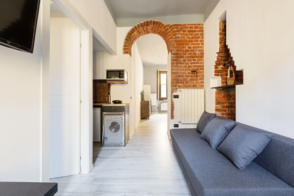 A modern living room with a grey sofa, a TV mounted on the wall, a brick fireplace, and a kitchen area in the background.