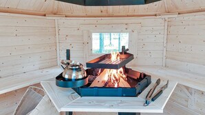 Interior de cabana de madeira com lareira central, uma janela e uma mesa com chaleira e utensílios.