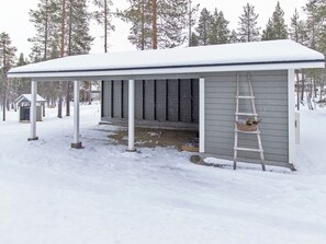 Un espace extérieur abrité, avec une échelle en bois appuyée contre un mur, entouré d’un sol et d’arbres recouverts de neige.