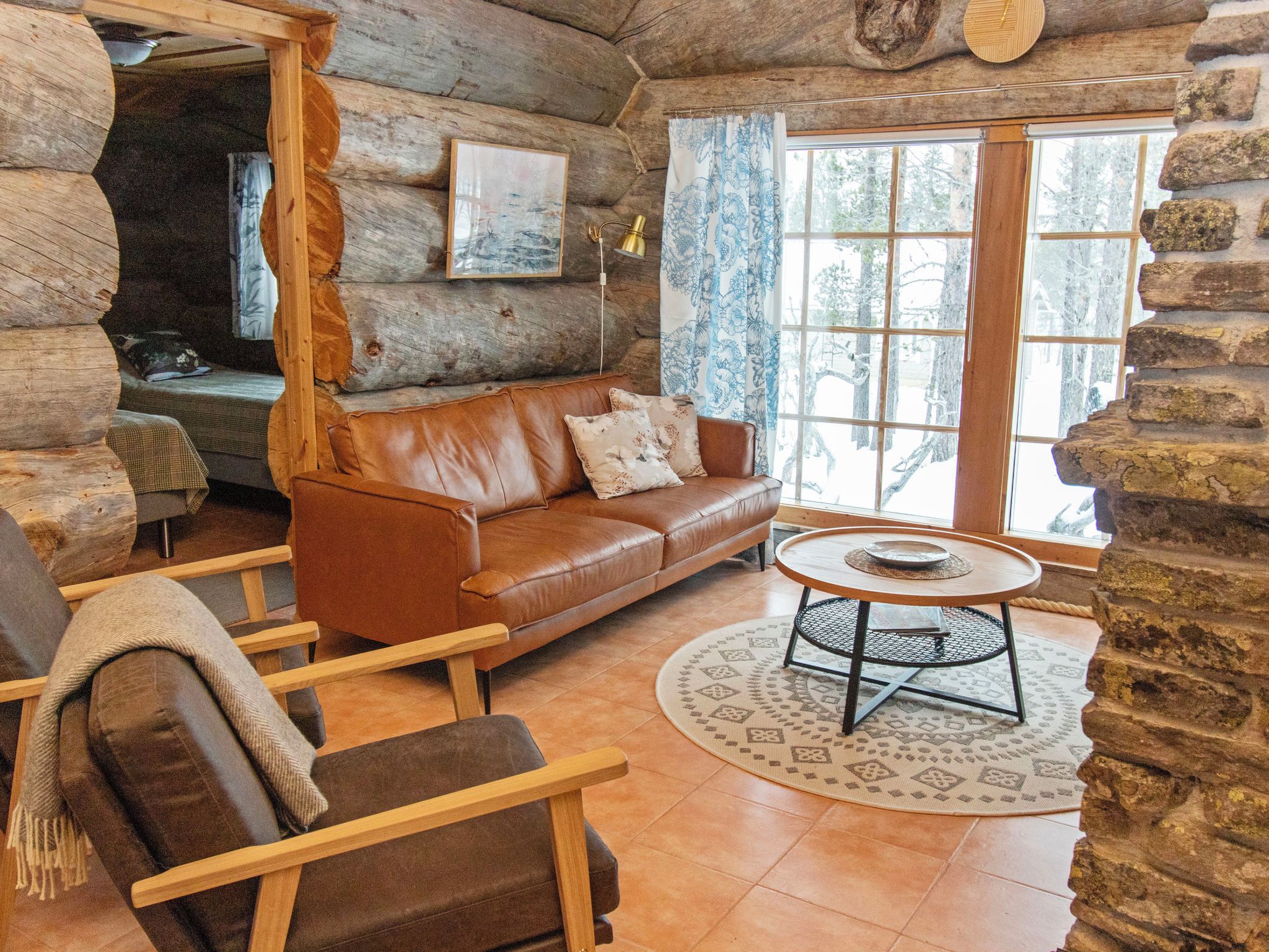 A log cabin interior with a leather sofa, a round table, and a view of the outdoors through a window.