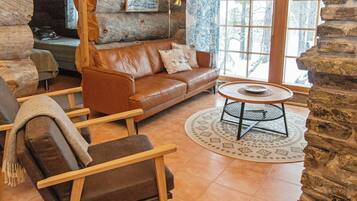 A log cabin interior with a leather sofa, a round table, and a view of the outdoors through a window.