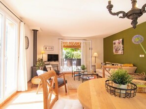 Una sala acogedora con una mesa de comedor de madera, un sofá y un televisor. Hay un ventanal con cortinas, un candelabro y una pared verde con un cuadro de flores.