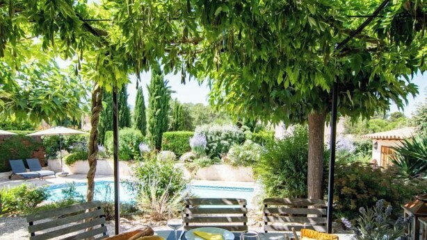 An outdoor dining area with a table set for two, surrounded by lush greenery and a pool.