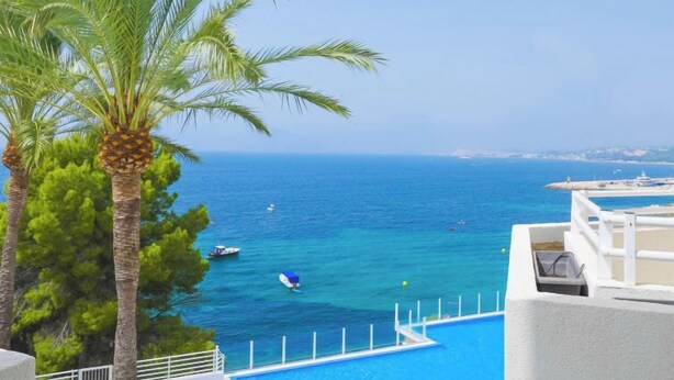 A poolside view with a clear blue sea, palm trees, and a distant shoreline. - Panorama by Interhome (Altea)