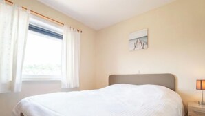 A bedroom with a bed, a nightstand, a lamp, a window with curtains, and a painting on the wall. - Green Garden VX 105 with pool view by Interhome (De Haan)