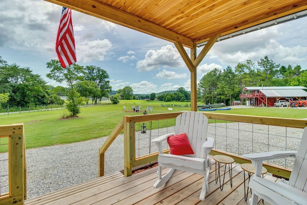 A wooden deck with white Adirondack chairs and a red pillow, overlooking a grassy area with a red barn and a blue tractor. - Norfork Studio by White River: Ideal for Fishing! (Norfork)