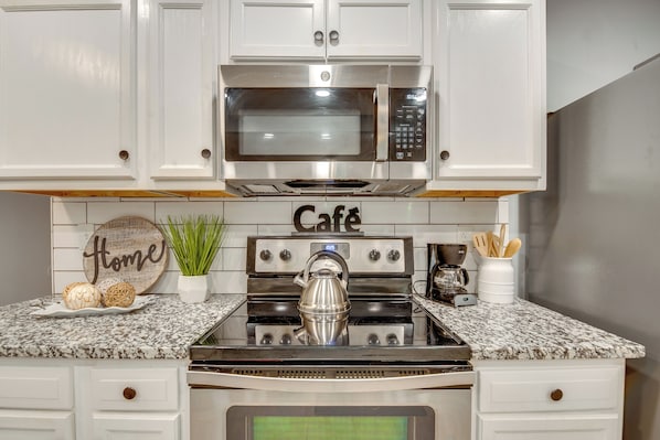 Una cocina con gabinetes blancos, un microondas de acero inoxidable, una estufa y una cafetera.