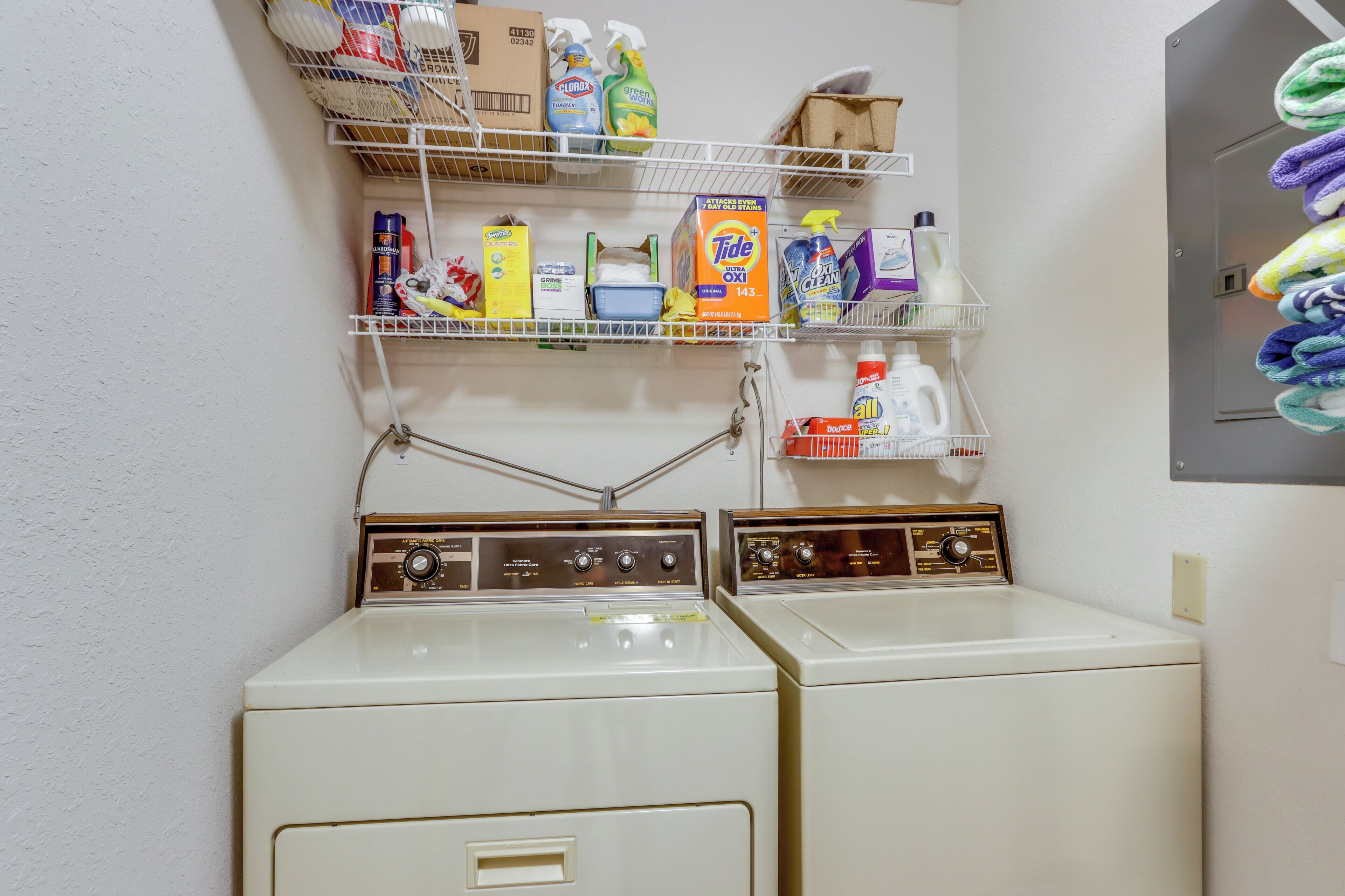 A laundry area with two white washing machines and shelves above containing cleaning supplies.