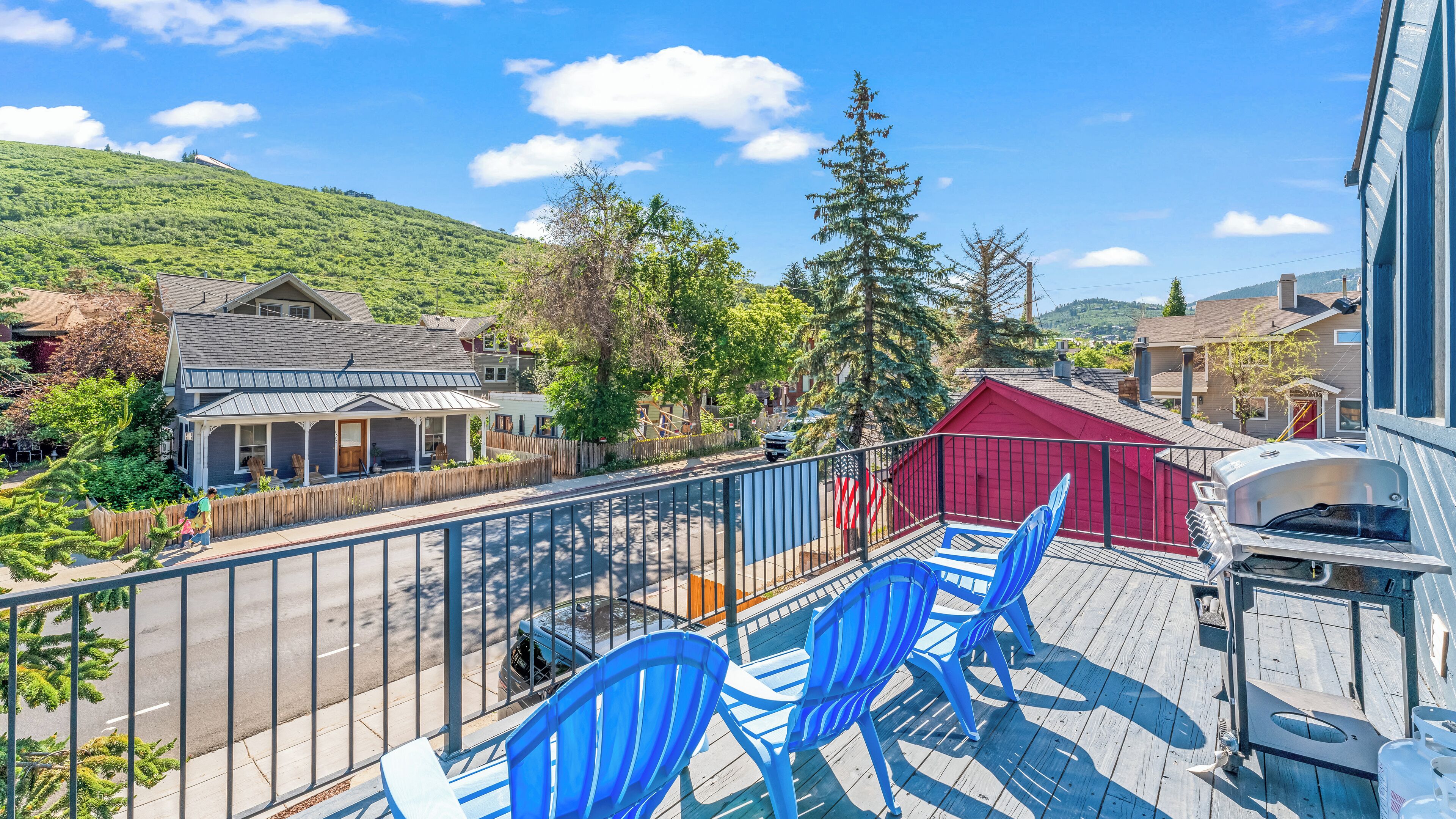 Una terraza con sillas azules, una parrilla y vistas a una zona residencial con casas y una colina.
