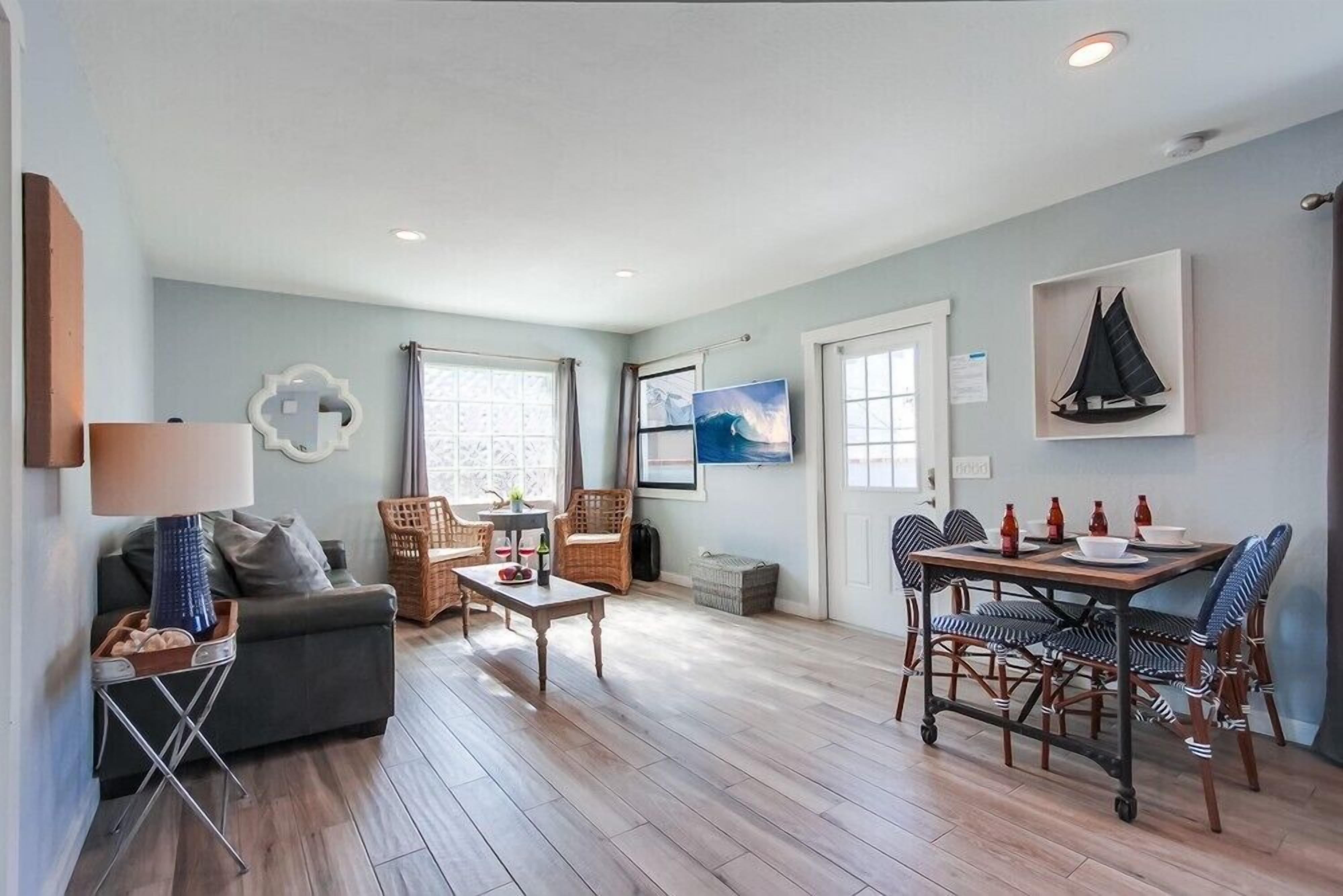 A cozy living room with a sofa, coffee table, and dining table set up. There are wicker chairs, a lamp, and artwork on the walls.
