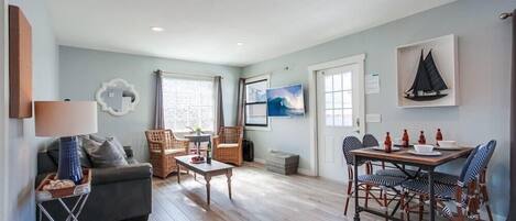 A cozy living room with a sofa, coffee table, and dining table set up. There are wicker chairs, a lamp, and artwork on the walls.