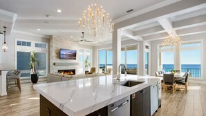 A spacious kitchen with a marble countertop, a built-in oven, and a large island with a sink. There is a fireplace with a TV mounted above it, and a dining area with a view of the ocean.