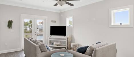 Una sala de estar con un sofá seccional color beige, una mesa de centro redonda, un televisor de pantalla plana sobre un mueble blanco y un ventilador de techo.