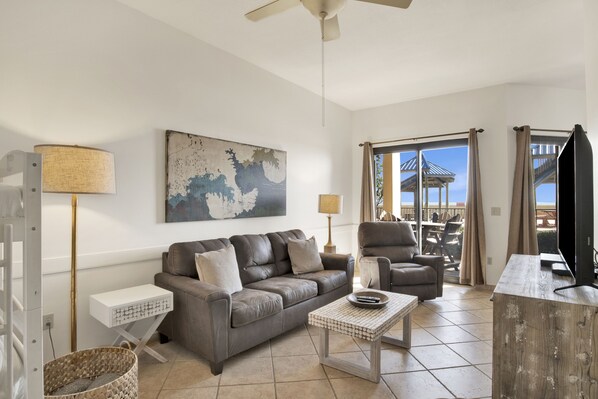 A living room with a sofa, armchairs, a coffee table, and a TV. There is a painting on the wall and a window with curtains. - Ground-Floor Beachfront 1BR + Bunk Alcove @ Phoenix V • Sleeps 6 • Pool, Beach & Splash Pad (Orange Beach)