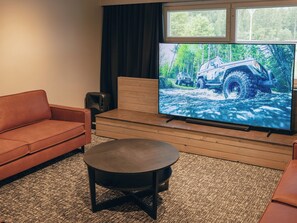 Un salon doté d’un grand téléviseur à écran plat qui diffuse des images d’un véhicule, d’un canapé en cuir marron, d’une table basse en bois et d’un sol recouvert d’une moquette.