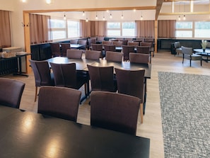 Uma sala de jantar ampla, com várias mesas e cadeiras, grandes janelas e um teto alto com vigas aparentes.