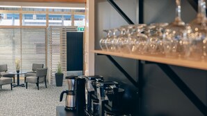 Un café moderne doté de machines à expresso et d’une vitrine réfrigérée. On y trouve des coins salon avec des chaises et une petite table agrémentée d’une plante.