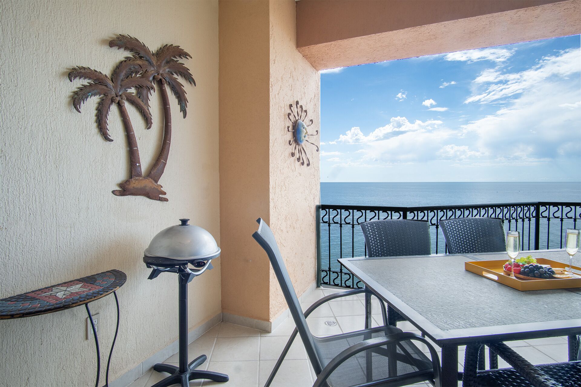 Een balkon met een tafel en stoelen, een decoratieve metalen palmboom en uitzicht op zee.
