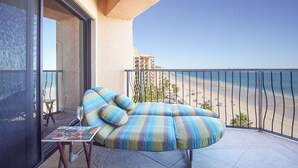 Un balcón con zona de estar y cojines a rayas, una pequeña mesa con una copa de vino, y vistas a una playa y edificios de hotel.