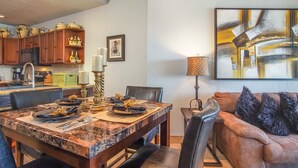 Un comedor con una mesa de mármol, sillas y un sofá. Se observa una cocina al fondo con armarios de madera y una cafetera.