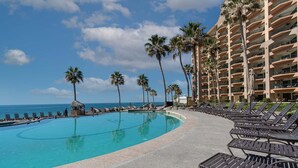 Piscina de un resort con sillas de descanso, palmeras y vista al océano.
