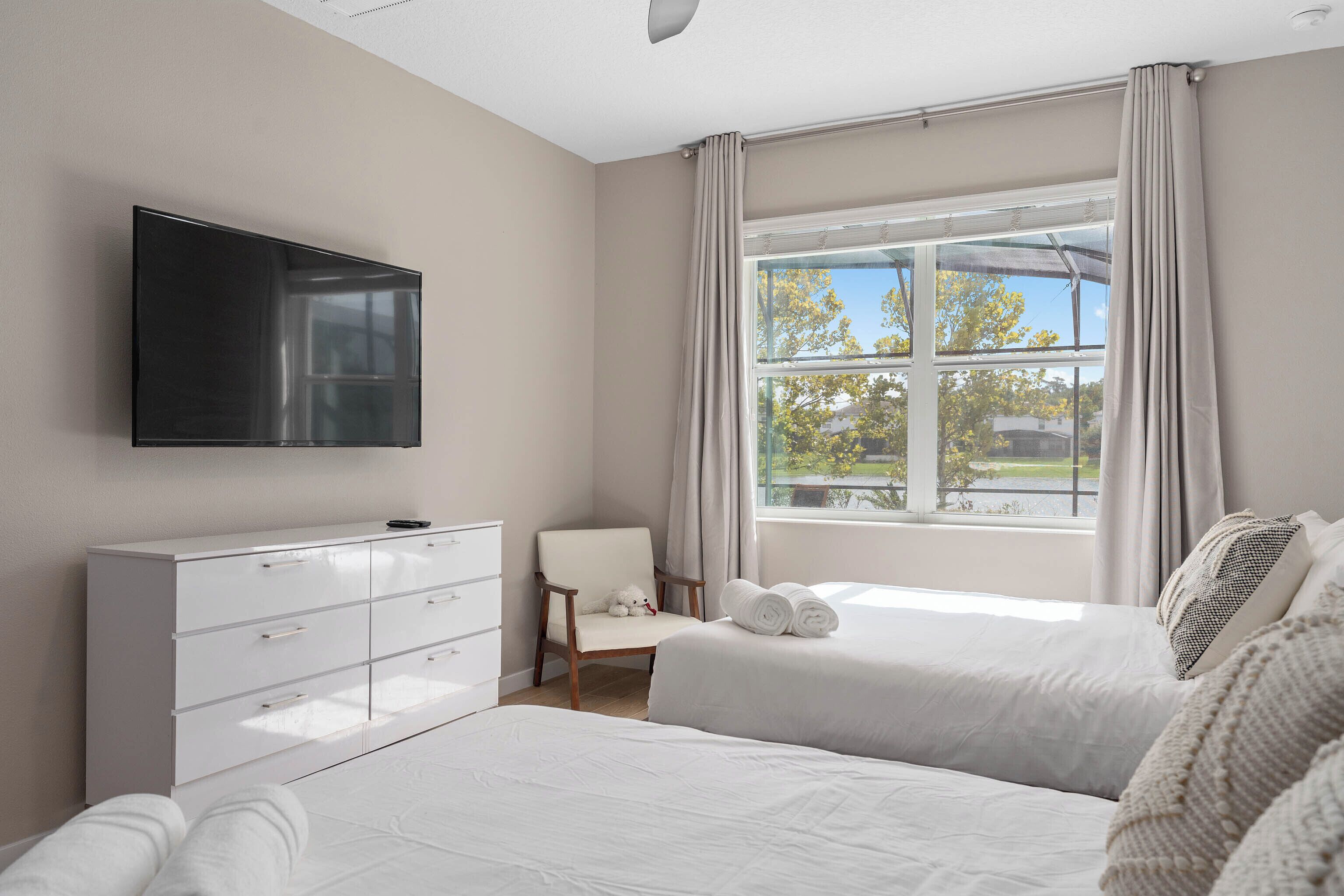A bedroom with a bed, a TV mounted on the wall, a dresser, and a window with curtains.