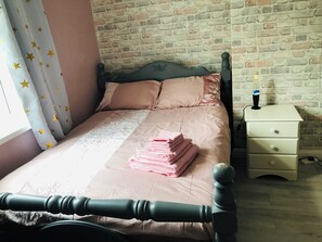 A neatly made bed with pink bedding, a nightstand with drawers, and a brick wall background. - Lovely 3-bed Charming Apartment in Portsmouth (Portsmouth)
