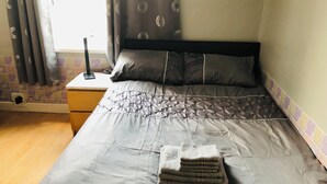 A neatly made bed with a grey duvet, a stack of folded towels, and a bedside table with a lamp.