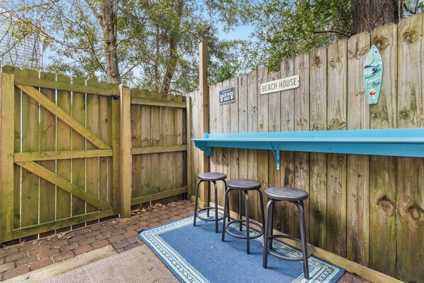 A wooden fence with a sign reading "Beach House" and a surfboard decoration. There are three metal barstools in front of a blue counter.