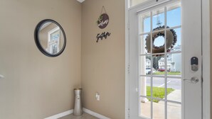 A hallway with a beige wall, a round mirror, a 'welcome' sign, a 'Gather' sign, a potted plant, and a door with a wreath. - Beach Sand House, Sleep 8, Swimming Pool, Gated E (Gulfport)