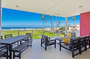 A balcony with a view of the beach, outdoor seating, and palm trees.