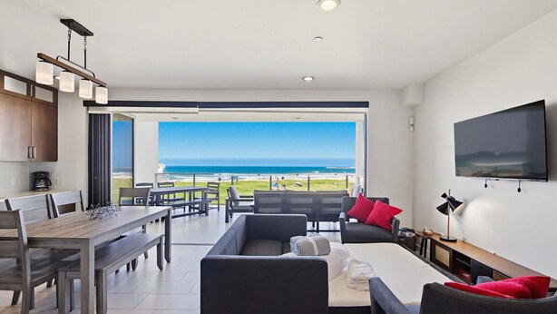 A modern living room with a dining area, a sofa set, a TV mounted on the wall, and a large window offering a view of the ocean. - Stylish Ocean Front Condo with Great Views (San Diego)
