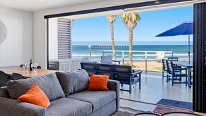 Una sala moderna con un ventanal grande con vista a la playa, un comedor con mesa y sillas, y un sofá con cojines.