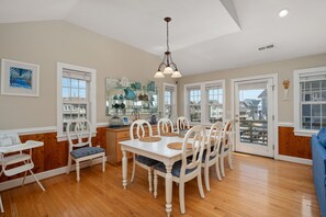 A dining area with a white table and chairs, a wooden floor, and a view of the outside. - Bella Vita: Pool, hot tub, volleyball/basketball, rec room, easy beach access! (Nags Head)