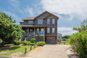 A two-story wooden building with a porch, a garage, and a clear sky. - Bella Vita: Pool, hot tub, volleyball/basketball, rec room, easy beach access! (Nags Head)