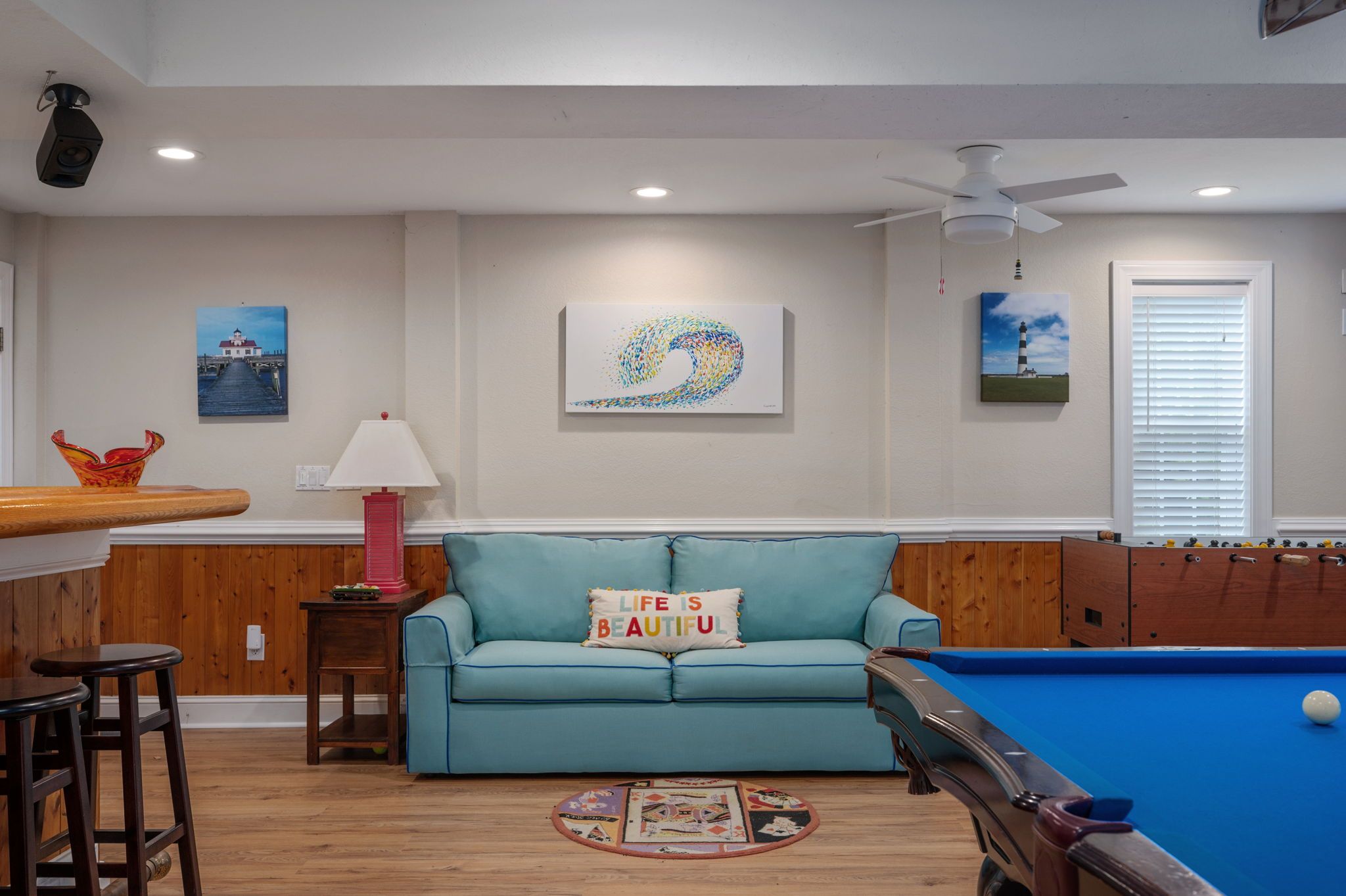 Un salon avec une table de billard, un canapé bleu avec un coussin, une lampe et des œuvres d’art accrochées aux murs.