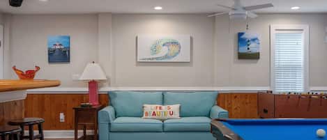 Un salon avec une table de billard, un canapé bleu avec un coussin, une lampe et des œuvres d’art accrochées aux murs.