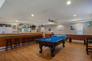 Een pooltafel in een ruimte met houten vloeren, een bar met krukken en een plafondventilator.