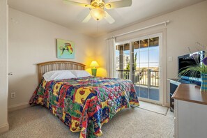 A bedroom with a colorful quilt, a ceiling fan, a window with curtains, and a view of a balcony.