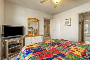 Une chambre à coucher avec une télévision posée sur un meuble en bois, une commode, un miroir, une porte et une couverture colorée sur le lit.