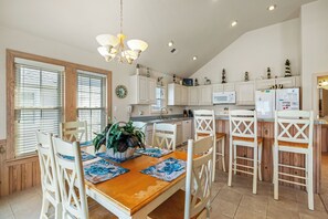 A kitchen with white cabinets, a dining table with chairs, and a chandelier. - Carolina Dreamin: Semi-oceanfront, pool, hot tub, ocean views, elevator! (Kill Devil Hills)