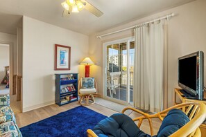 A living room with a blue rug, a bookshelf, a chair, a television, and a view of the outdoors through a sliding glass door. - Carolina Dreamin: Semi-oceanfront, pool, hot tub, ocean views, elevator! (Kill Devil Hills)