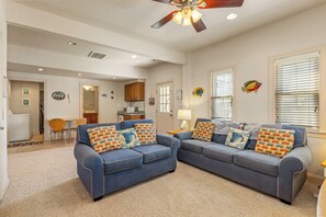 A living room with two blue sofas, a ceiling fan, and a kitchen in the background.
