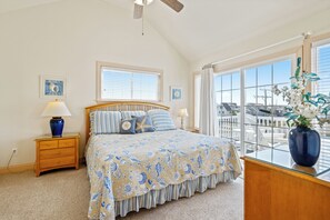 A bedroom with a bed, a nightstand, a lamp, a window with blinds, and a view of the outside. - Ocean Escape: Oceanside, pool, hot tub, pool table, easy beach access (Kill Devil Hills)